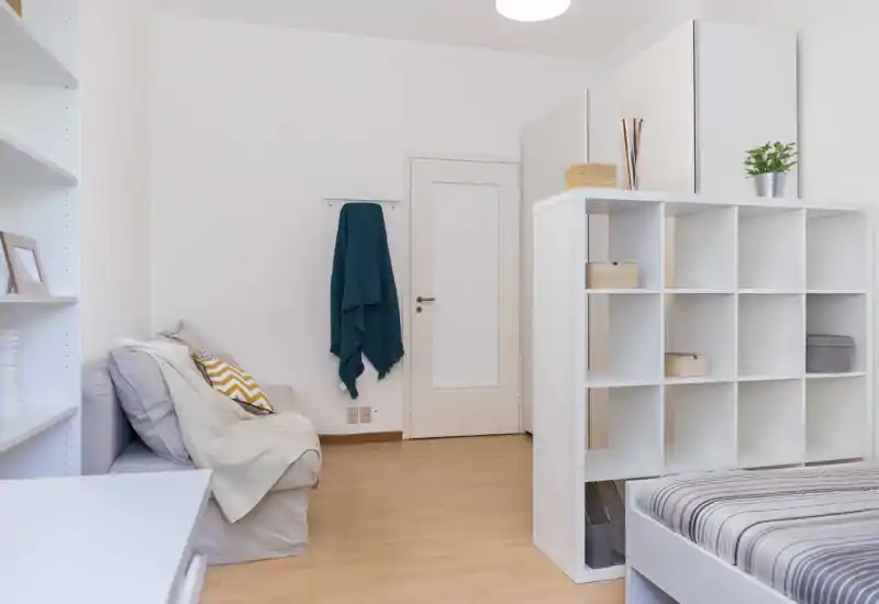 Bright, minimal bedroom corner with a partially visible bed, white shelving unit used as a room divider, soft sofa and cozy cushions — tidy and modern studio-style layout.