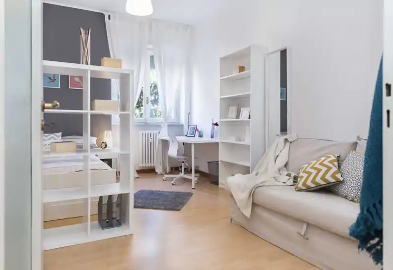 Bright and airy bedroom with a comfortable bed partially visible behind a white shelving unit, a desk by the window and a cozy sofa with patterned cushions — ideal for a single occupant or a stylish studio layout.