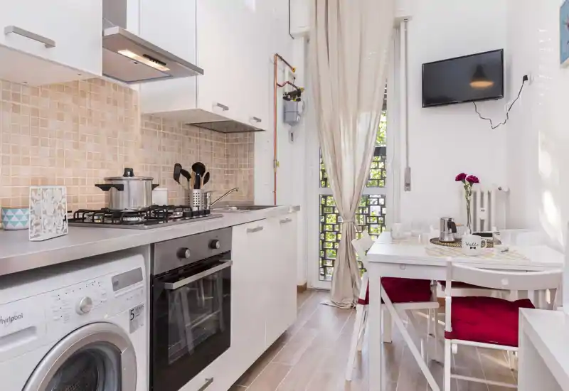 Compact modern kitchen with integrated oven, gas hob and washing machine; small dining table by a bright window creates a cozy breakfast corner.