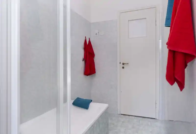 Bagno pulito con piastrelle chiare, vasca con parete doccia in vetro e asciugamani colorati a contrasto.