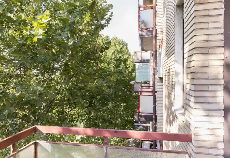 Balcone soleggiato con alberi rigogliosi e facciate degli edifici vicini; ideale per un caffè mattutino o rilassarsi all'aperto.