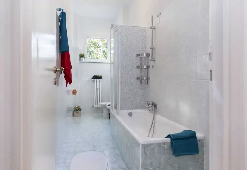Bagno luminoso e pulito con vasca e box doccia parziale, piastrelle su pareti e pavimento, termosifone e luce naturale dalla finestra.