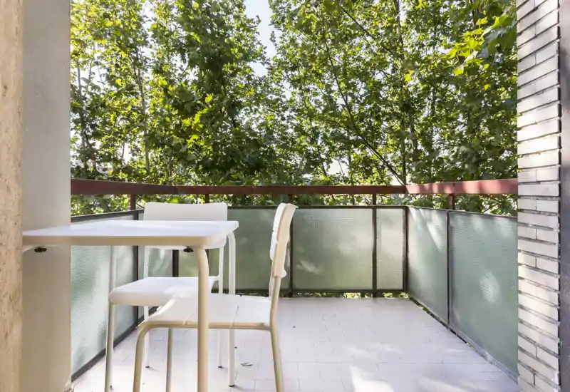 Sunny private balcony with a small table and two chairs, framed by green trees — ideal for morning coffee or relaxing outdoors.