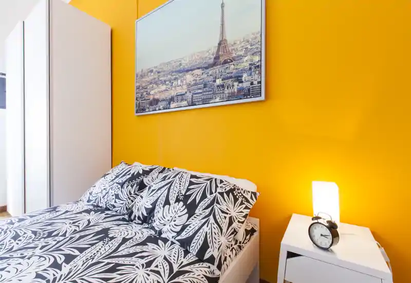 Camera da letto moderna e luminosa con parete arancione, biancheria a fantasia e comodino con lampada e sveglia — ambiente accogliente e ben arredato, ideale per un annuncio.