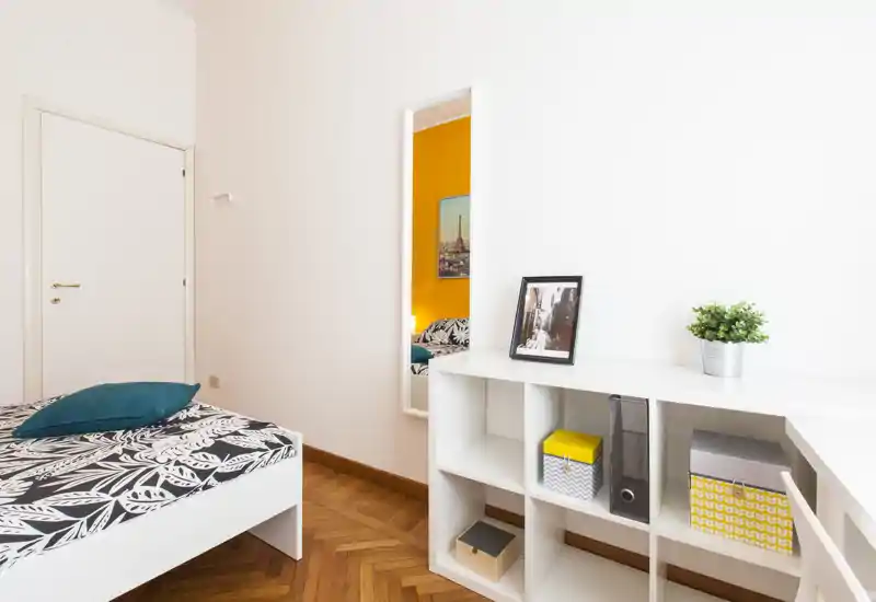 Camera singola luminosa e moderna con biancheria a motivo, cuscino turchese e scaffalatura bianca; arredi minimali e pavimento in legno caldo donano un ambiente fresco e accogliente.