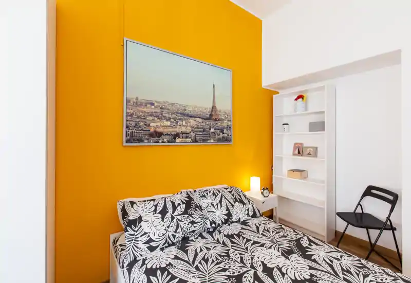 Chambre moderne et lumineuse avec lit double à motifs devant un mur d'accent orange, lampe de chevet et étagère — un espace chaleureux et bien décoré, idéal pour la location courte durée.