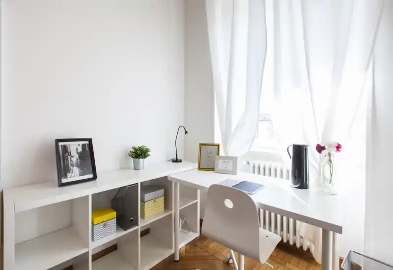 Coin bureau lumineux et minimaliste avec bureau blanc, chaise et étagères près d'une fenêtre ensoleillée — espace de travail propre et moderne, idéal pour le télétravail.
