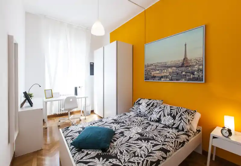 Camera da letto moderna e luminosa con letto matrimoniale, biancheria a fantasia e parete arancione vivace. Ampia finestra che garantisce molta luce naturale e una scrivania per lavorare.