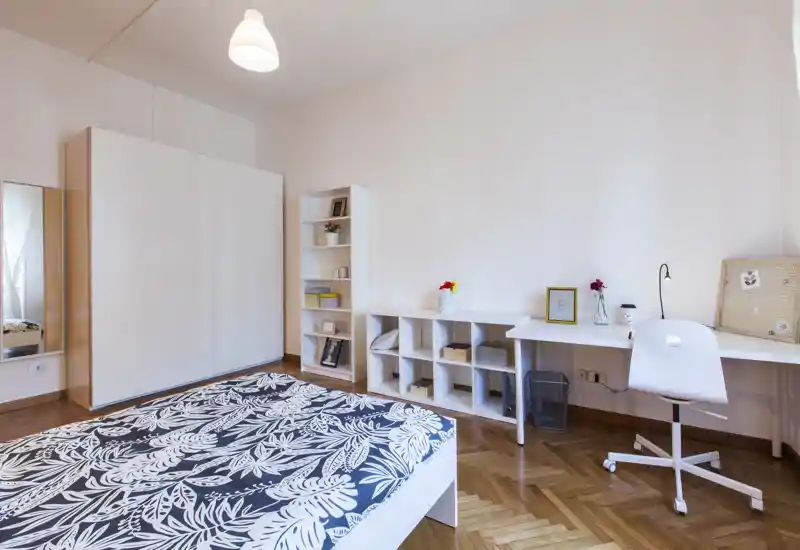 Chambre lumineuse et soignée avec un lit double à motif au premier plan, grand placard blanc, étagères et un long bureau avec chaise — style moderne et belle lumière naturelle.