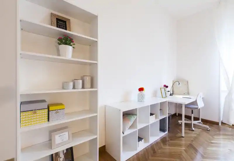 Espace séjour/bureau lumineux et minimaliste avec étagères blanches, rangement en cubes et petit bureau près d'une fenêtre ensoleillée — propre, moderne et bien rangé.