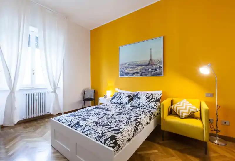 Camera da letto luminosa e moderna con letto matrimoniale in biancheria a fantasia, parete d'accento gialla e una poltrona accogliente vicino alla lampada da terra. Ampia finestra con tende leggere, pavimento in parquet e quadro decorativo.