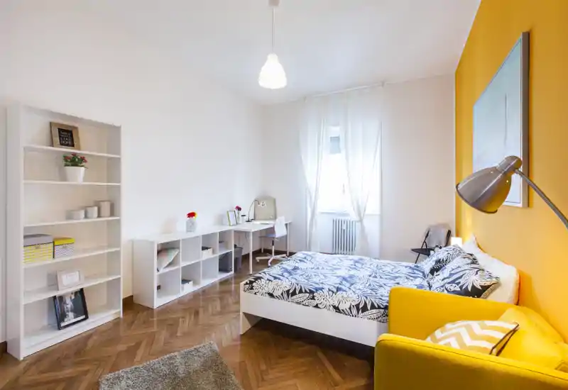 Camera da letto luminosa e moderna con letto matrimoniale, biancheria decorata e parete color giallo caldo; comprende scrivania, scaffalatura e una poltrona accogliente accanto al letto.