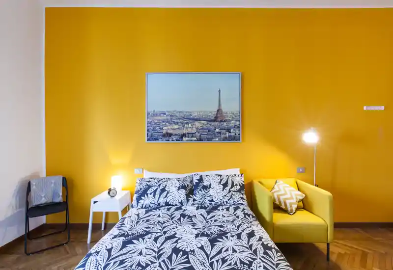 Camera da letto luminosa e di stile con letto matrimoniale con biancheria decorata, parete gialla, comodino e poltrona — accogliente e ben illuminata.