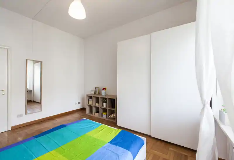 Camera da letto luminosa e minimal con letto matrimoniale con biancheria colorata, un grande armadio bianco e una piccola libreria a cubi. La stanza ha luce naturale dalla finestra e parquet.