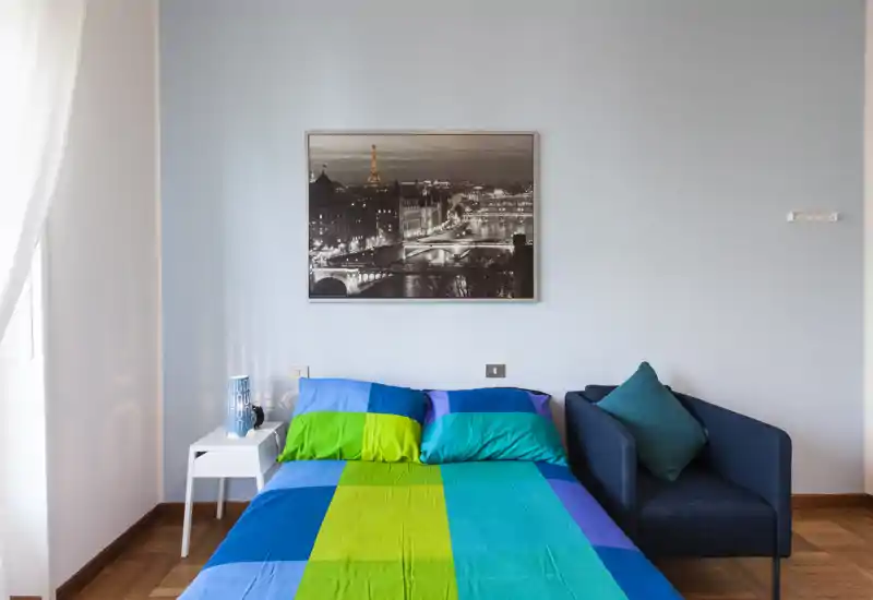 Camera da letto moderna e luminosa con letto matrimoniale dai colori vivaci, comodino con lampada e una poltrona. Arredamento pulito e minimale con una stampa della città sopra il letto.