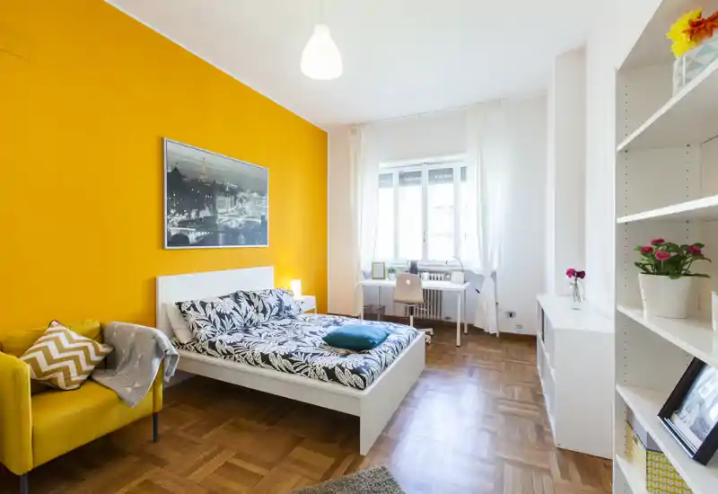 Camera da letto luminosa e dal design moderno con parete gialla, letto matrimoniale con biancheria a fantasia, poltrona comoda e scrivania vicino alla finestra — perfetta per riposo e smart working.