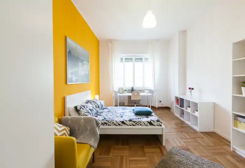 Camera da letto luminosa e di stile con letto matrimoniale davanti a una parete gialla accogliente, scrivania vicino alla finestra e scaffali ordinati — ideale per un soggiorno confortevole.