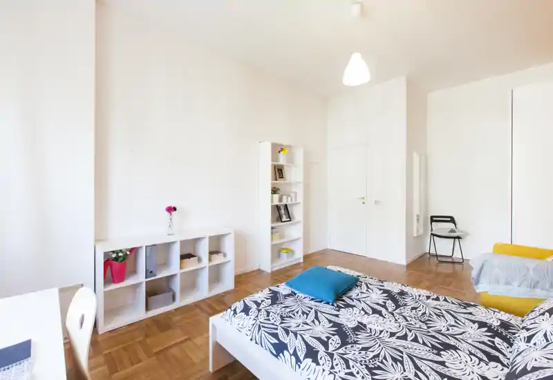 Camera da letto luminosa e moderna con letto matrimoniale in primo piano, scaffalatura bianca e parquet in legno chiaro. Stile minimal pulito, tessili accoglienti e un angolo salotto comodo, perfetto per annunci.