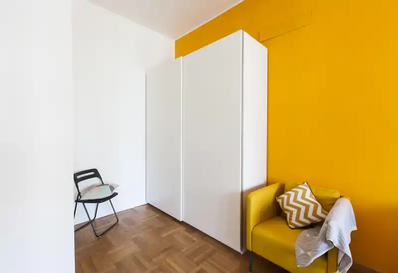 Coin de chambre avec un mur d'accent jaune vif, une armoire blanche et un fauteuil jaune cosy avec coussin et plaid.