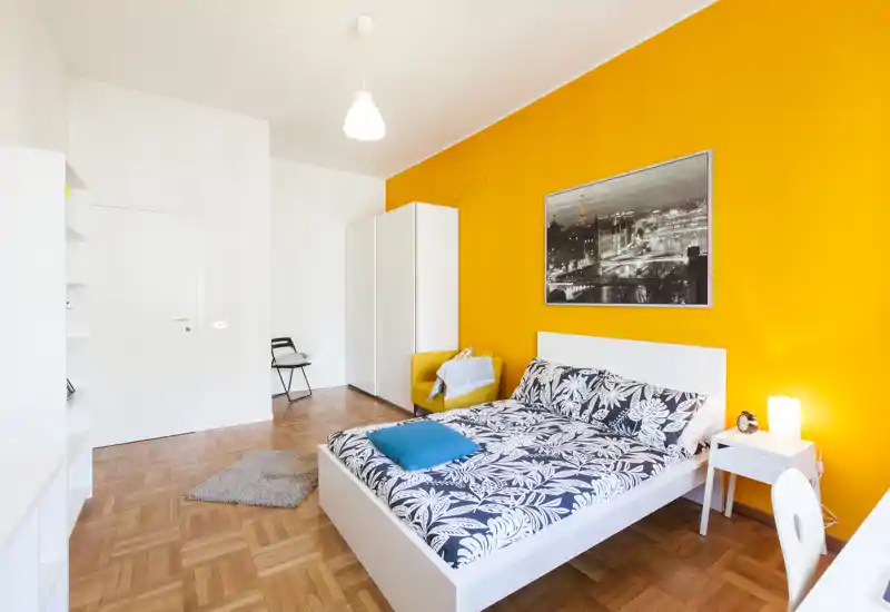 Camera da letto luminosa e moderna con letto bianco, biancheria con fantasia e una parete arancione vivace. Pavimento in parquet, armadio, comodino con lampada e quadro decorativo rendono l’ambiente accogliente.