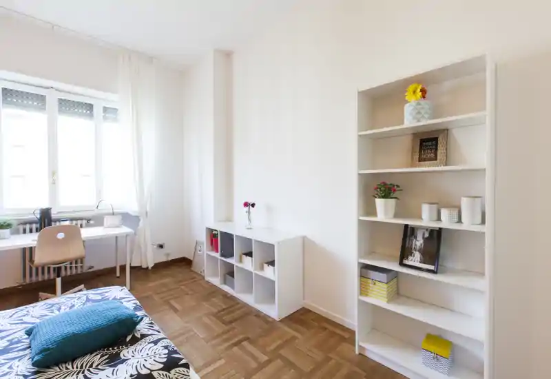 Chambre lumineuse et minimaliste avec lit à motif, parquet, bureau près de la fenêtre et étagères de rangement — idéale pour une personne seule ou un étudiant.