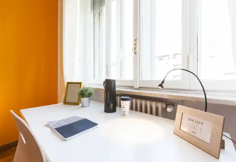 Espace de travail lumineux et moderne près d'une grande fenêtre avec bureau blanc, lampe, cadres décoratifs et petite plante — idéal pour lire ou travailler à la lumière naturelle.