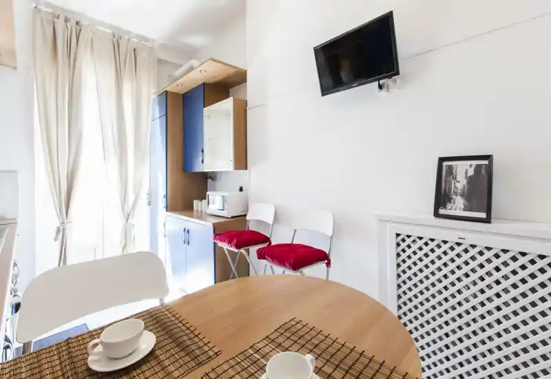 Coin repas lumineux avec table ronde en bois, chaises blanches et coussins rouges; grande luminosité grâce aux rideaux et petite kitchenette en arrière-plan.