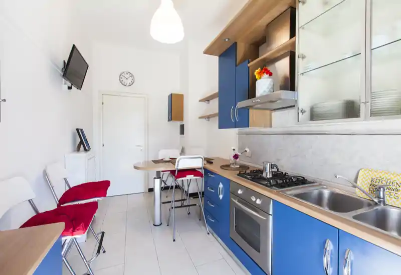 Cucina moderna e luminosa con pensili blu, piano cottura a gas, forno incassato e doppio lavello. Bancone con sgabelli e cuscini rossi che rendono l'ambiente accogliente.