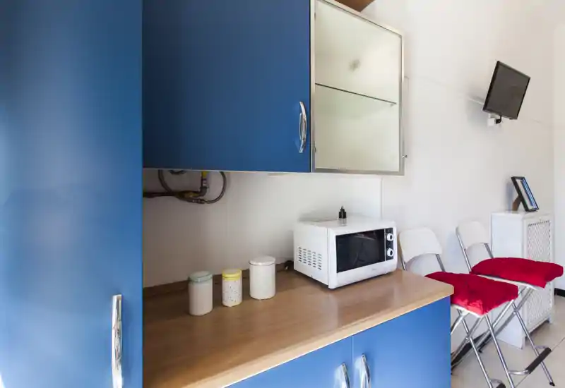 Coin cuisine compact avec des placards bleus, un micro-ondes sur le plan de travail et trois boîtes décoratives; deux chaises pliantes avec coussins rouges et une télévision murale sont visibles.