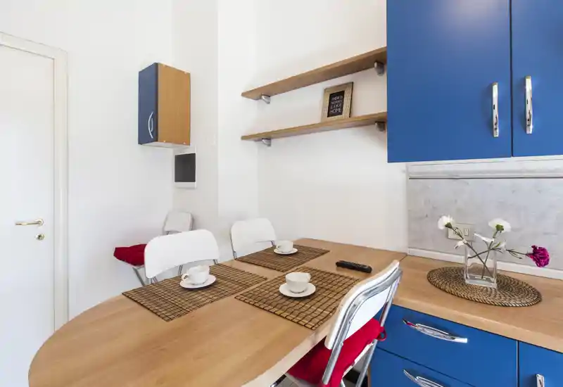 Cucina compatta e luminosa con piano in legno, pensili blu e un piccolo angolo pranzo apparecchiato con tazze e tovagliette — perfetta per pasti accoglienti.