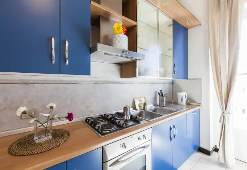 Cucina moderna compatta con ante blu, piano cottura a gas, forno integrato e lavello in acciaio. La luce naturale e i dettagli (fiori, tovaglietta) creano un ambiente luminoso e accogliente.