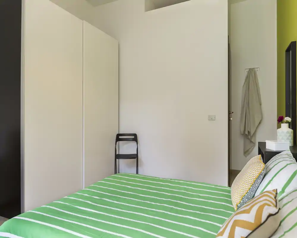 Dormitorio luminoso y ordenado con colcha verde a rayas, cojines decorativos y mobiliario mínimo: un espacio para dormir fresco y acogedor.