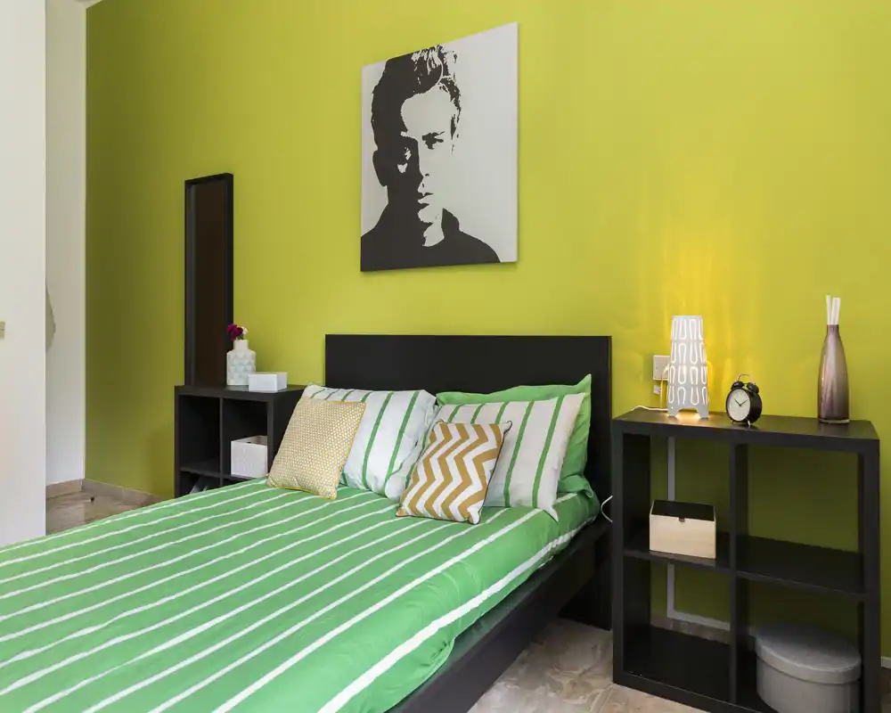 Dormitorio moderno y luminoso con cama de rayas verdes, cojines decorativos y estanterías a juego junto a la mesita; líneas limpias y un cuadro contemporáneo crean un ambiente fresco y con estilo.