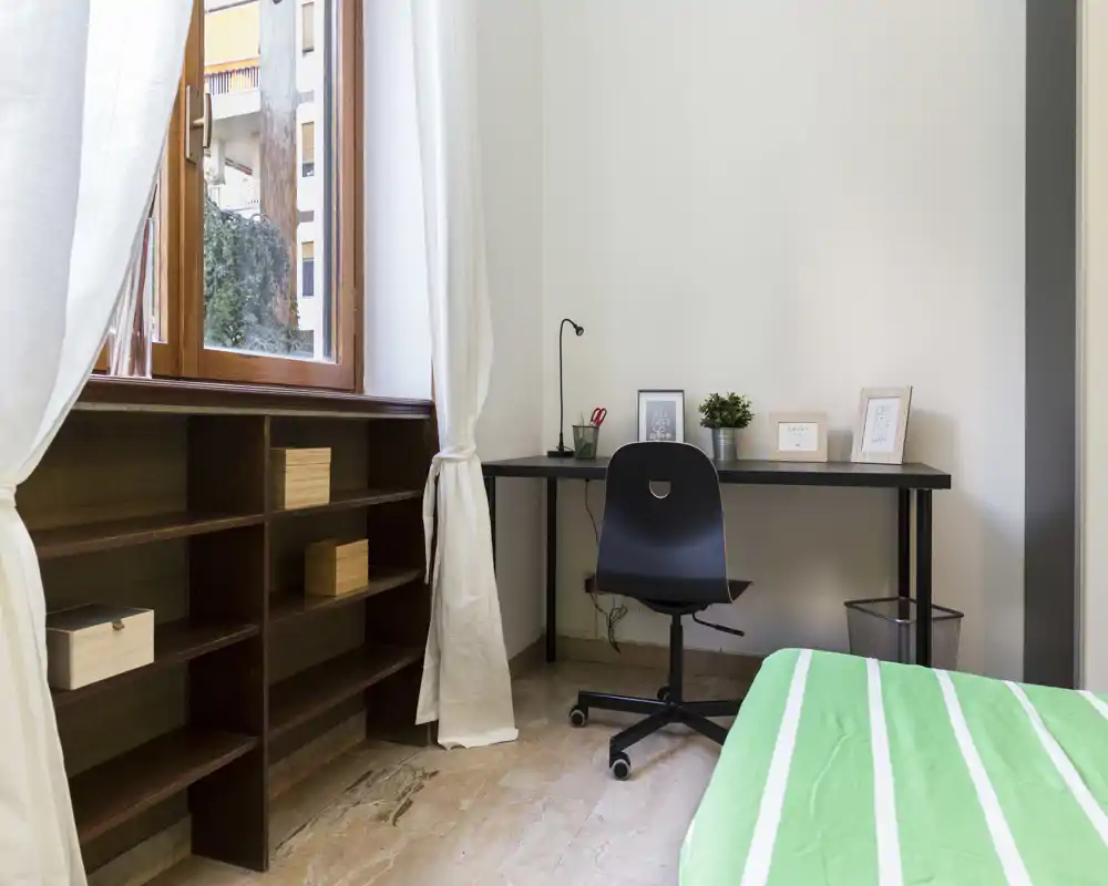 Rincón de dormitorio luminoso y ordenado con cama individual, escritorio y estanterías; la luz natural de la gran ventana hace agradable el área de estudio.