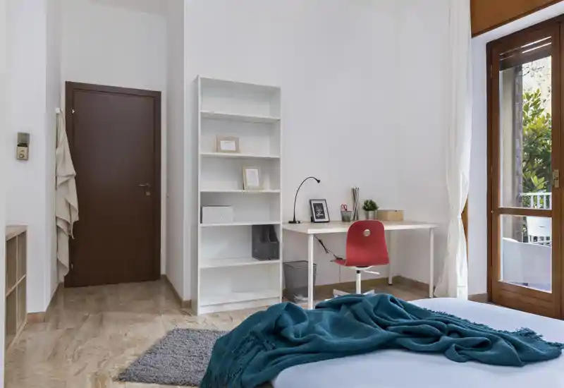 Bright, modern bedroom corner with a neatly made bed, textured throw and a small workspace by the balcony offering natural light.