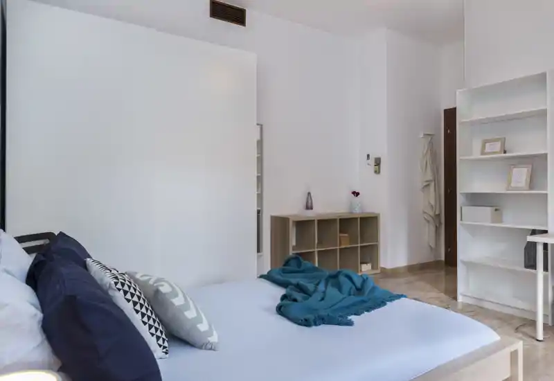 Camera da letto moderna e luminosa con letto ben rifatto, cuscini decorativi e una coperta turchese; scaffalature e armadio semplici che creano un ambiente ordinato e arioso, perfetto per gli annunci.
