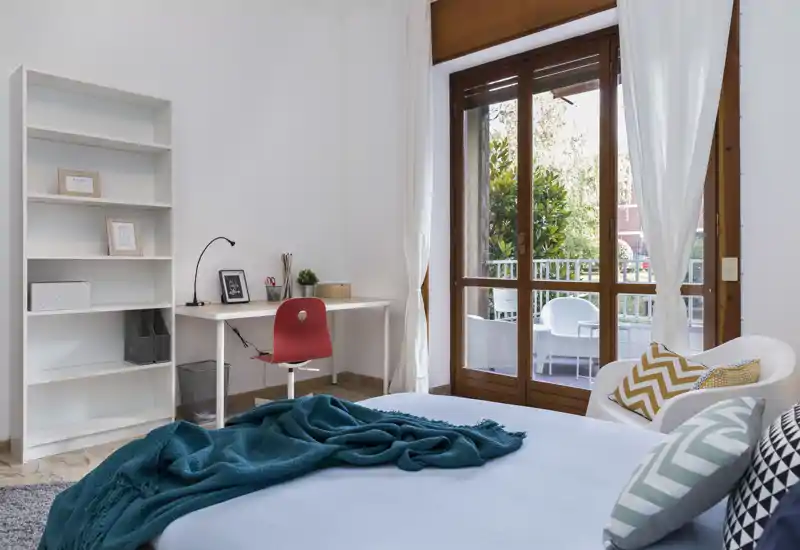 Camera da letto luminosa e curata, con letto ben rifatto, cuscini decorativi e plaid turchese, scrivania funzionale e porta finestra verso il balcone che illumina l'ambiente.