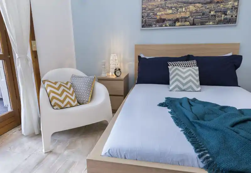 Camera da letto luminosa e moderna con struttura letto in legno, cuscini decorativi e plaid turchese; una poltrona e un comodino con lampada rendono l’ambiente accogliente.