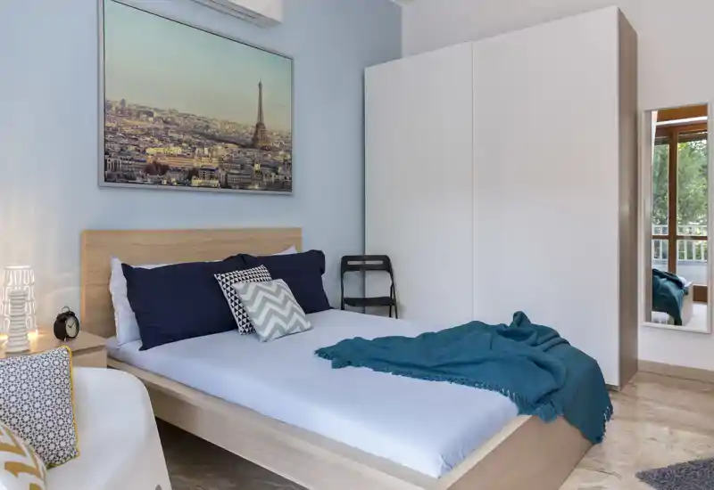 Camera da letto moderna e luminosa con struttura letto in legno, cuscini navy e decorativi e plaid turchese; grande armadio e stampa di Parigi per stile e spazio.