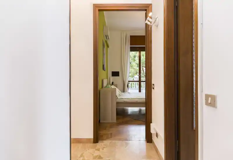 A bright, neatly staged bedroom seen through a hallway doorway, with a double bed, bedside table and a balcony window letting in natural light.