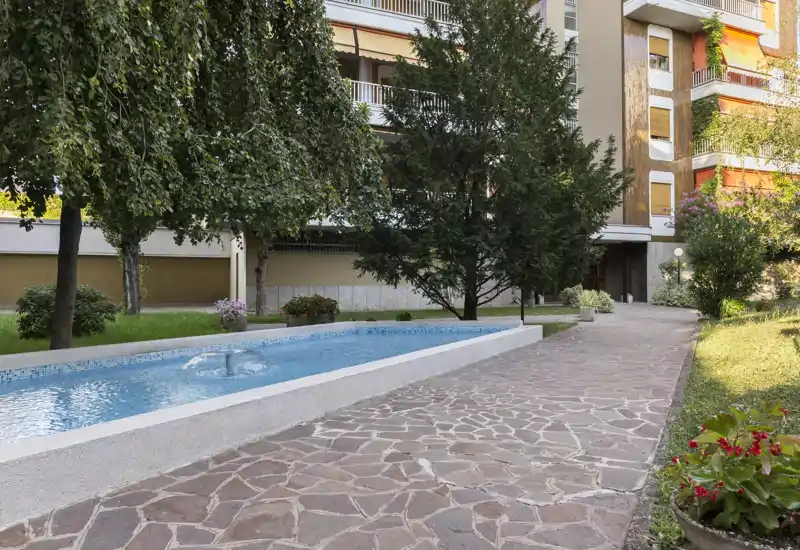 Sunny courtyard with a decorative rectangular fountain, paved walkway and landscaped greenery in front of an apartment building — ideal for relaxing outdoors.