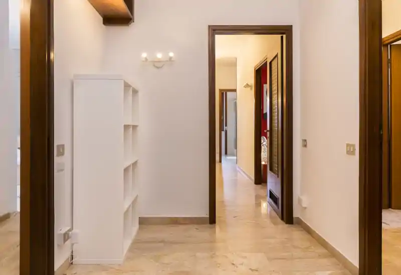 Bright, tiled entrance corridor with warm wooden door frames and a slim white shelving unit — clean, well-lit passage connecting rooms.