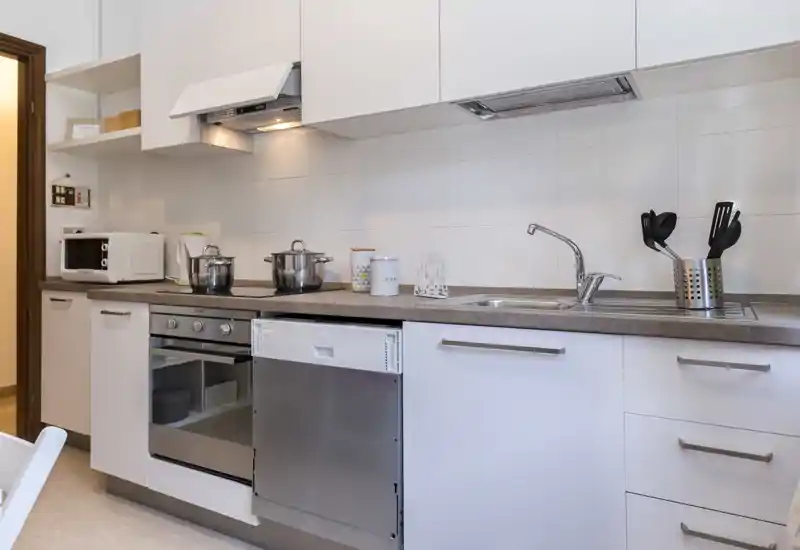 Modern, well-lit kitchen with integrated oven, dishwasher and sink; clean white cabinets and useful counter space with utensils and cookware.