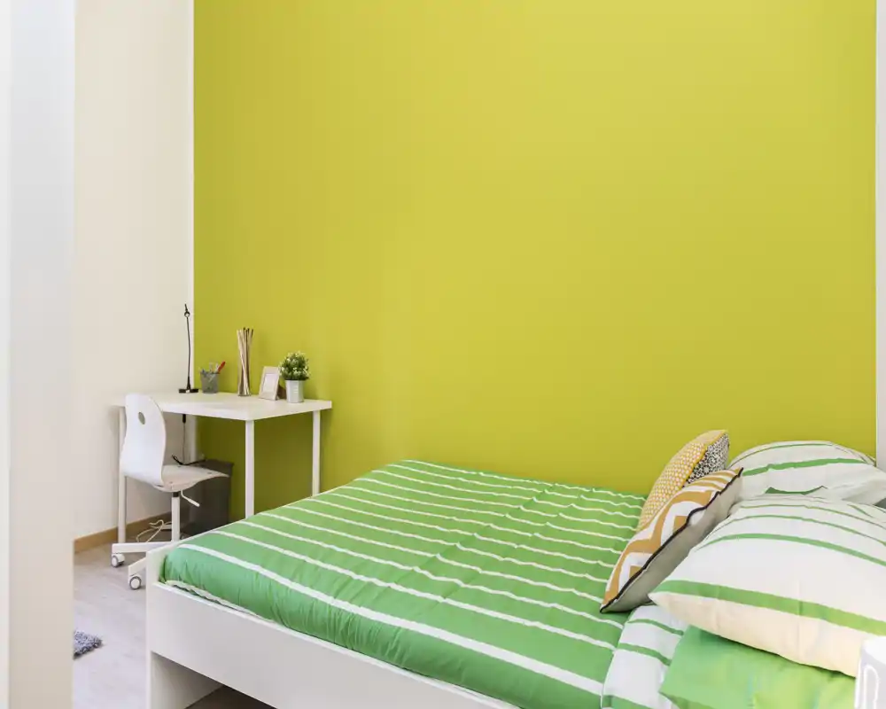 Camera da letto moderna e luminosa con letto a righe verdi e cuscini coordinati; una piccola scrivania bianca e una sedia sono contro la parete color lime, creando uno spazio fresco e accogliente adatto a riposo e studio.