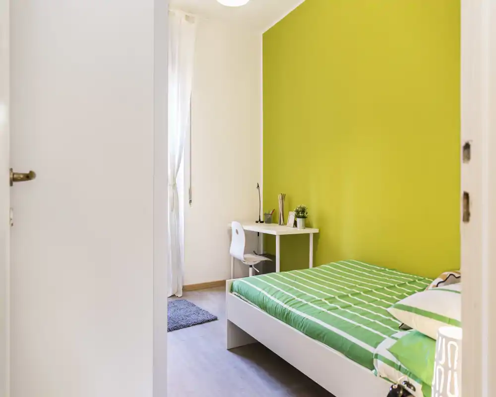Camera da letto luminosa con parete verde a contrasto e letto singolo ben rifatto; una scrivania vicino alla finestra offre un pratico angolo studio.