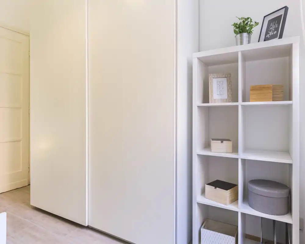Area camera luminosa e minimale con ampi armadi a muro e una libreria organizzata con scatole decorativi e una pianta.