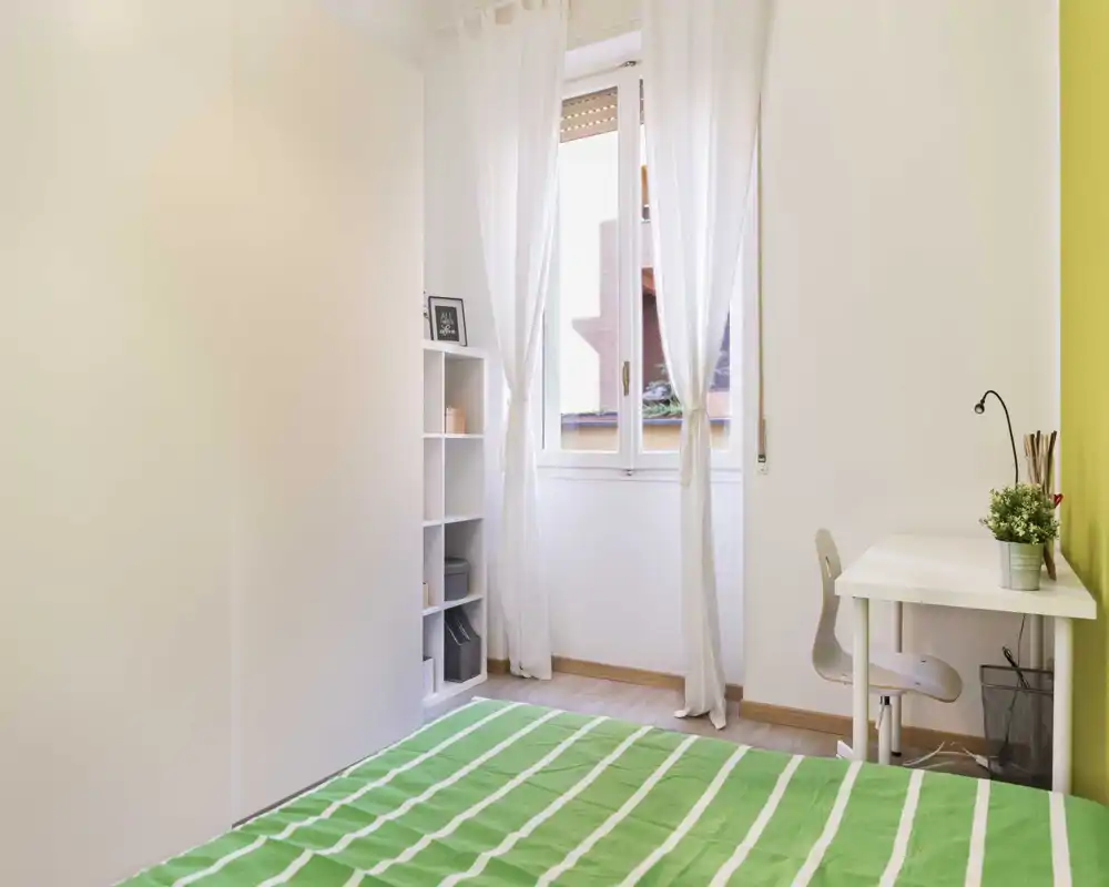 Camera da letto luminosa e minimale con copriletto a righe verdi, mobili bianchi e una scrivania vicino alla finestra — spazio pulito e arioso ideale per una persona o uno studente.