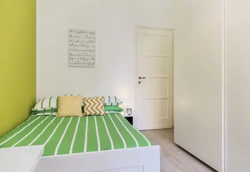 Camera da letto luminosa e ordinata con letto a righe verdi, cuscini decorativi e una lampada da comodino semplice; pareti e armadio bianchi per un aspetto moderno e fresco.