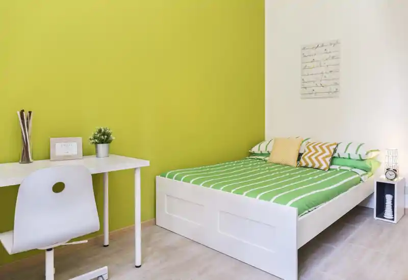 Camera da letto luminosa e moderna con letto bianco e biancheria verde a righe, cuscini decorativi e un piccolo comodino. Una scrivania bianca e una sedia sono posizionate contro una parete accentata verde, creando uno spazio accogliente e ordinato, ideale per studenti o soggiorni brevi.