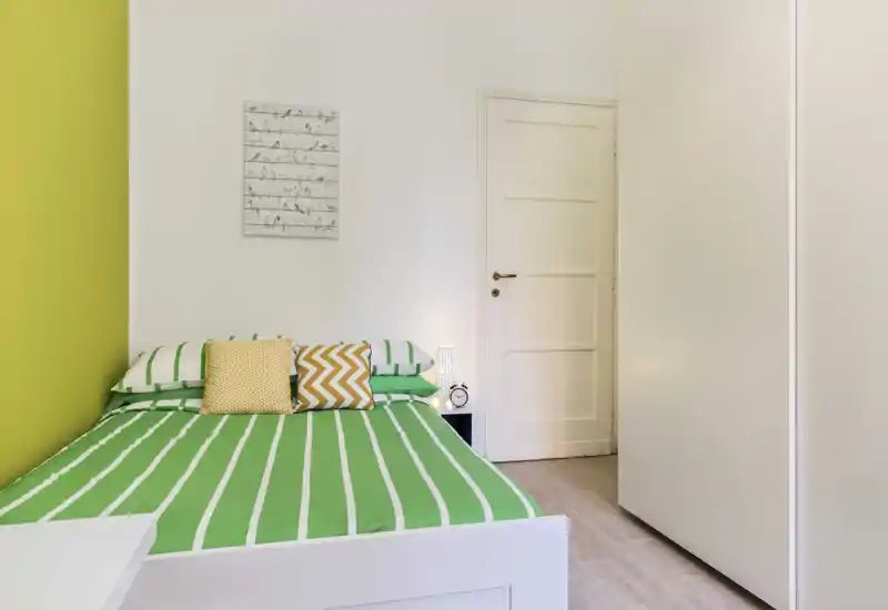 Chambre lumineuse et soignée avec un lit à cadre blanc habillé d’une parure rayée verte et blanche, coussins jaunes décoratifs, lampe de chevet et armoire blanche — ambiance fraîche, moderne et accueillante.